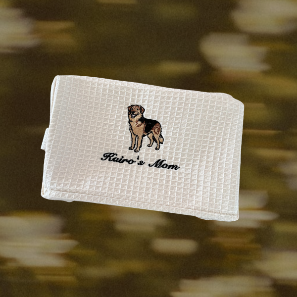 A white waffle-weave towel featuring an embroidered dog illustration and the text 'Rairo's Mom' in black script, shown against a blurred brown and gold background.