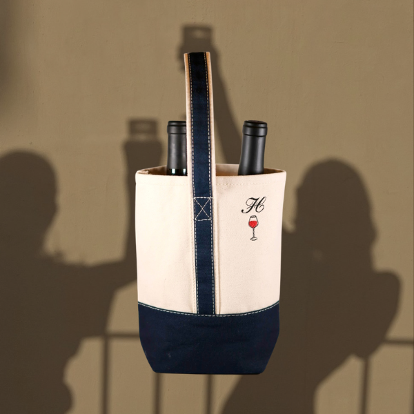 A canvas wine tote bag with navy blue trim and base, holding two wine bottles. The bag has a vertical embroidered monogram 'M' with a wine glass icon.